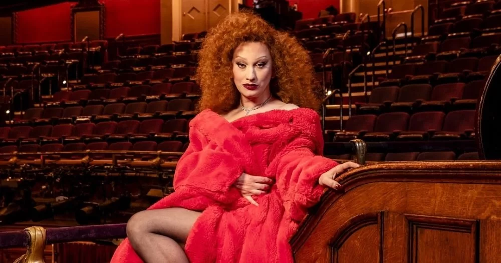 Jinkx Monsoon poses in an old theatre filled with red-upholstered seats. She wears a flowing red gown and heels. Her red hair is full of secrets.