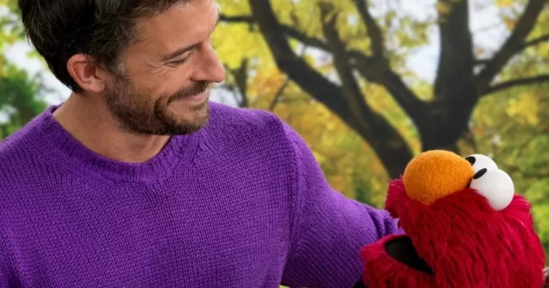 Johnathan Bailey appears on Sesame Street. Bailey and Elmo share a moment on a bench in a beautiful park surrounded by trees covered in leaves. Bailey wears a purple jumper.