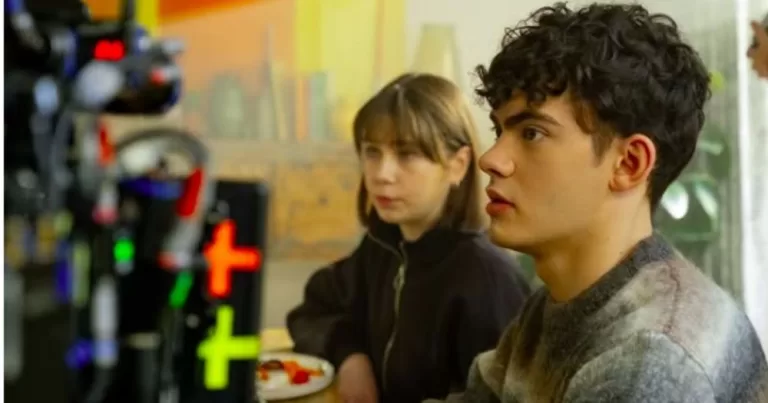 The image shows a behind the scenes of Hearstopper, with a camera on the left hand side facing character Charlie Spring, who is sitting next to his sister Tori, both wearing jumpers and staring off-camera. The image is an example of LGBTQ+ representation in children's film and TV.