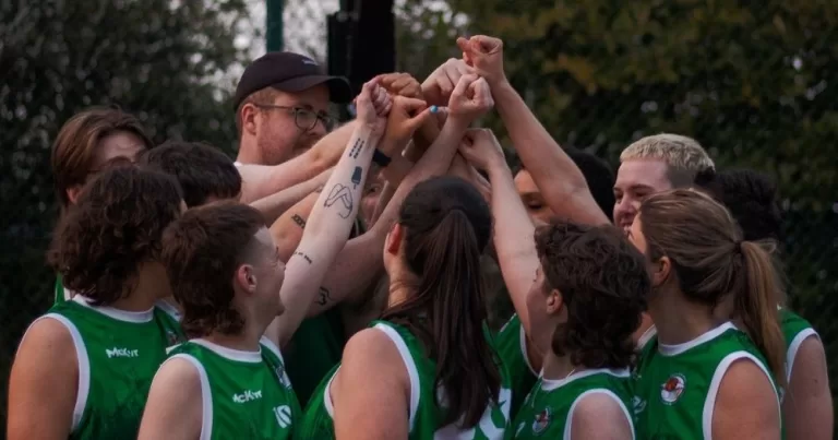 LGBTQ+ sports clubs in Ireland 2025. Shamrock Síoga, a queer basketball team wear green jerseys and hold their hands up in a huddle, in a basketball court.