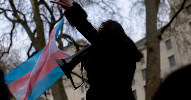Image shows a person holding a trans flag while talking into a speakerphone. This is being used to represent Transgender Day of Remembrance.