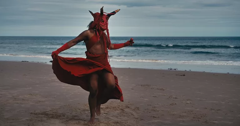 Screenshot from Out of Shadows, a film that will premiere on World AIDS Day 2025. In the photo, a person dancing on a beach wearing a red drape and mask.
