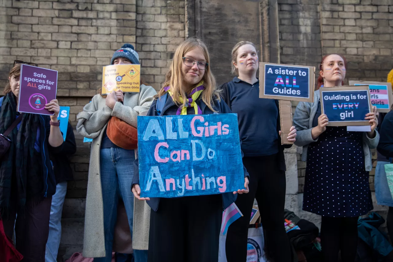 A girl wears a girl guide uniform and holds a sign that says All Girls Can Do Anything. She stand in front of other protestors holding signs.