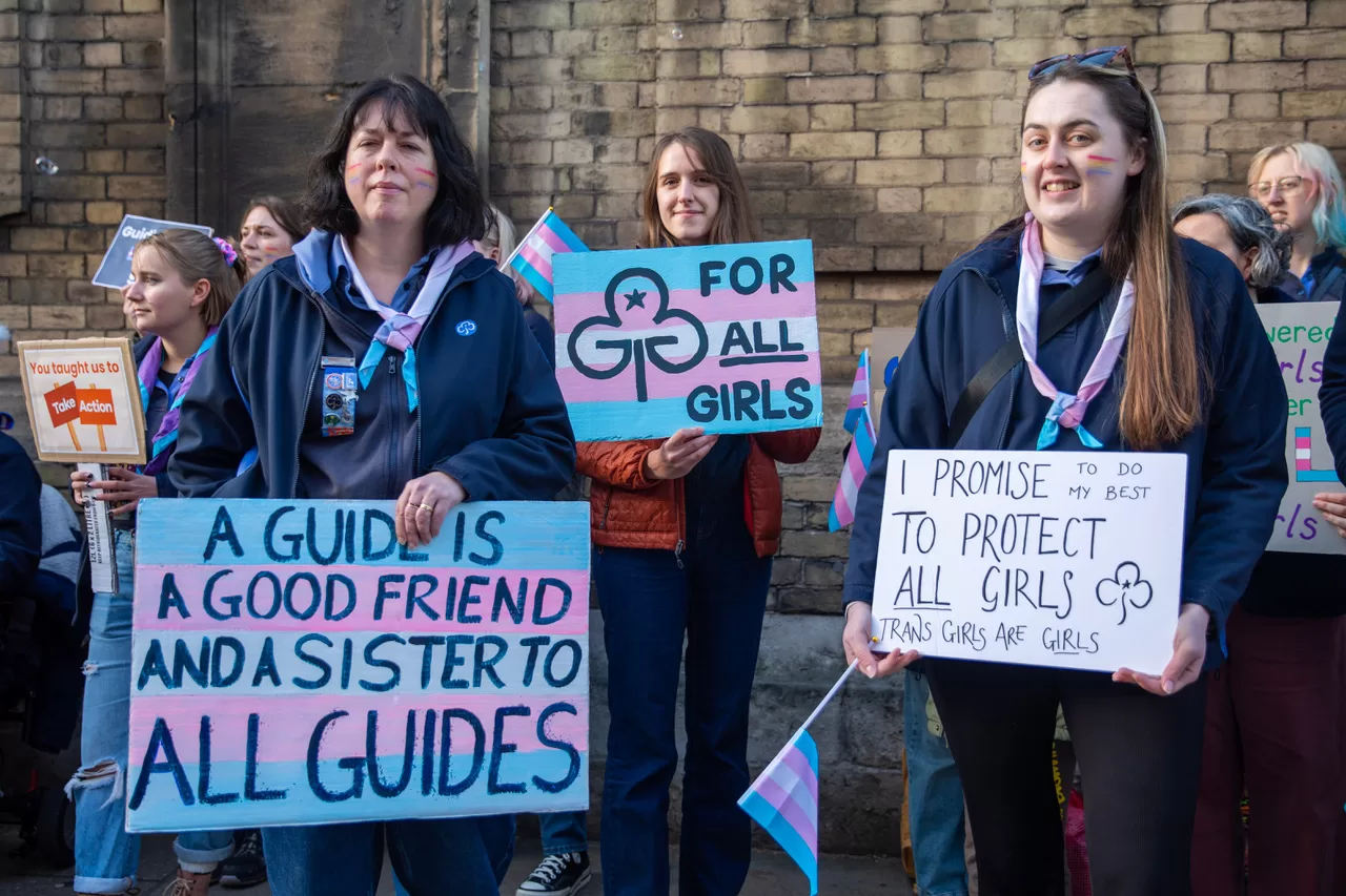 Protestors in Girl Guide uniforms hold trans flags and wear neckerchiefs in trans colours. They hold signs which say: A Guide is a good friend and sister to all guides, For all girls and I Promise to do my best to protect all girls, trans girls are girls.