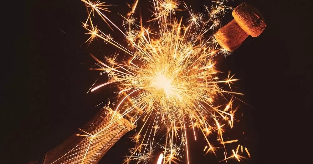 A firework goes off behind a bottle of champagne being opened.