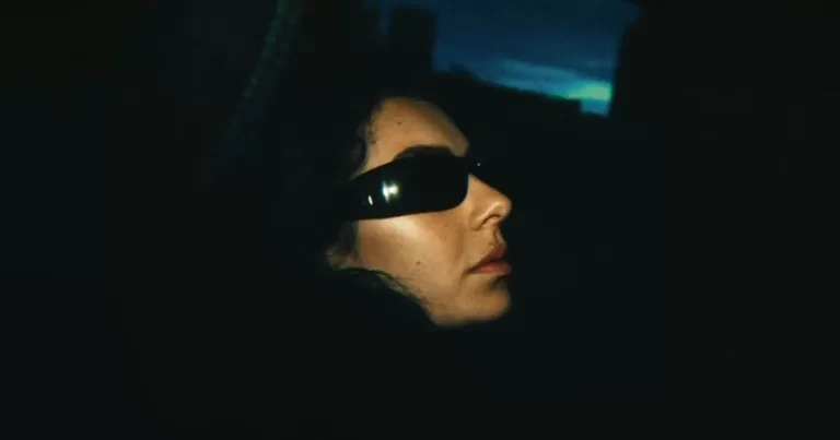 Trailer released for Charli XCX and A24's The Moment. A portrait of Charli with her signature sunglasses on in a dark abyss.
