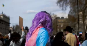 Image shows a person wrapped in a trans flag at a demonstration. This image accompanies an article about the National Gender Service.