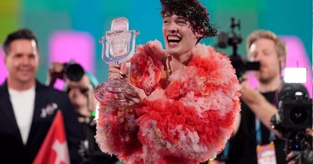 The image shows 2024 Eurovision winner Nemo, holding their trophy and smiling off camera. They are wearing a large pink and red fluffy coat, and the background show camera crew.