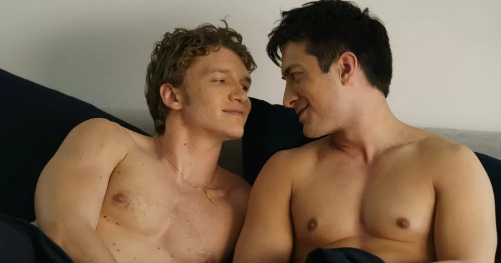 Heated Rivalry is finally coming to Ireland. The image shows Connor Storie and Hudson Williams gazing lovingly at each other while shirtless in bed.