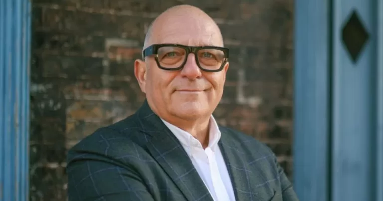 Photo of Hugh Wallace looking at the camera and smiling while wearing a grey suit and thick-framed glasses.