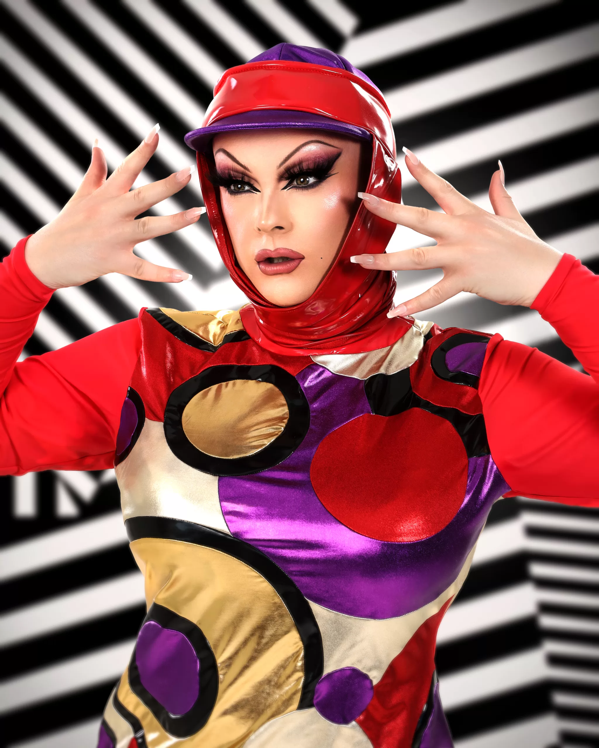 Bonnie Ann Clyde poses in a unique, patterned, futuristic outfit in front of a black and white stripy background.