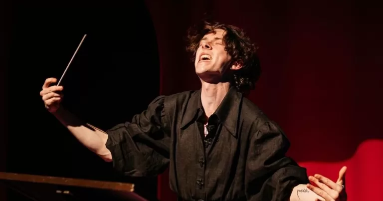 Disrupt Disability Arts Festival. A performance artist, Day Magee, emphatically conducts, wearing a black shirt, with mid length black curly hair, against a dark backdrop.
