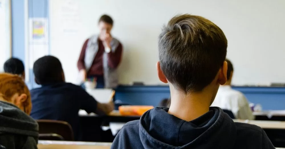School children sit in a classroom while a teacher is at a whiteboard.