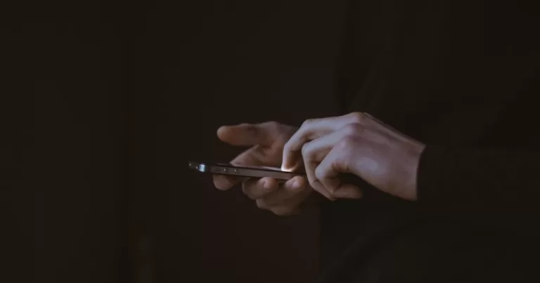 This article is about how to stay safe from sextortion on Grindr. In the photo, the hands of a person holding a phone in the dark.