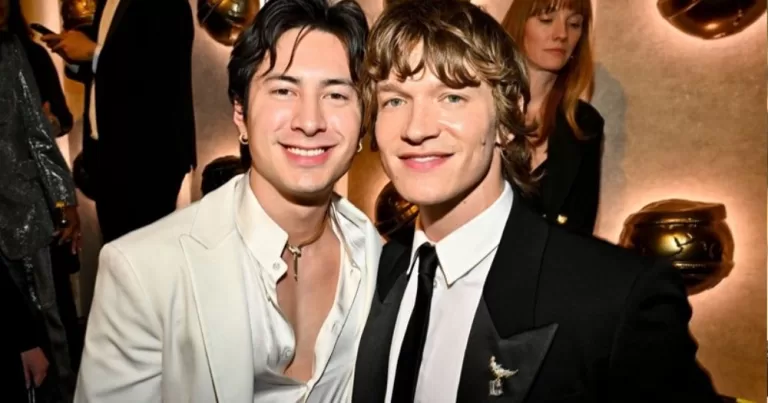 The image shows co-stars Hudson Williams (left) and Connor Storrie (right) on the red carpet for the Golden Globes. The Heated Rivalry actors are smiling at the camera, standing next to each other. Williams is wearing a white open suit, and Storrie is wearing a black suit jacket, white shirt, and black tie. The pair will carry a torch at the Winter Olympics soon.