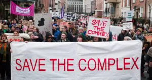 People protesting to save The Complex after the arts centre announced its closure. Marching in the streets, people are holding banners that read 'Save the Complex'.
