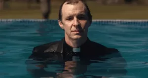 The image shows a still from the series These Sacred Vows, with Tom Vaughan-Lawlor pictured from the torso up in a swimming pool. He is wearing a black suit with a white priest's collar.