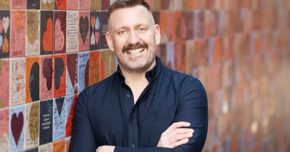The image shows new Belong To CEO Kieran O'Donovan, smiling at the camera with his arms folded. He is wearing a navy shirt and posing against a colourful background wall.