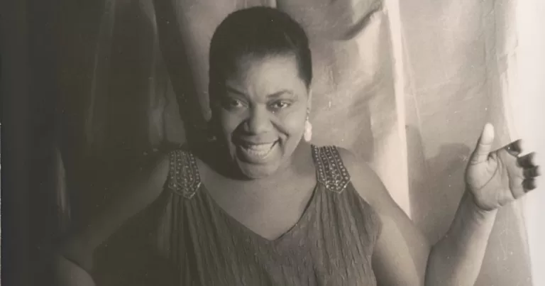 Blues singer Bessie Smith smiling at the camera while wearing an elegant dress in a black and white photo.
