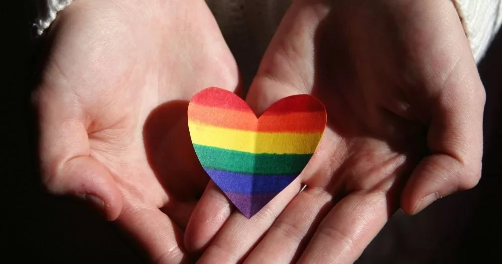 A person holds a rainbow-coloured paper heart in their hands. This image is being used to accompany an article about queer lent.