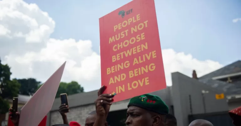 A red protest sign in Uganda reads "people must not choose between being alive". This article covers the arrest of two women in Uganda for kissing.