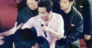 Leslie Cheung holds a microphone. He is surrounded by people.