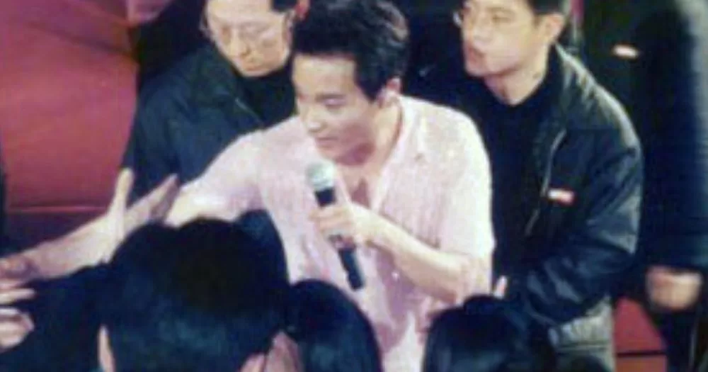 Leslie Cheung holds a microphone. He is surrounded by people.