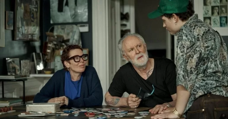 Colman, Lithgow, and Mason-Hyde in Jimpa. The three sit around a lovely wooden table. Colman wears incredibly chic chunky glasses.