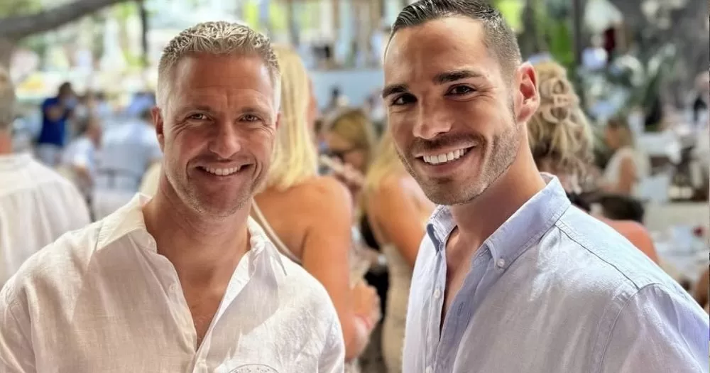 The image shows former F1 racer Ralf Schumacher on the left, and fiancé Étienne on the right. Both are smiling at the camera, and wearing light-coloured shirts.