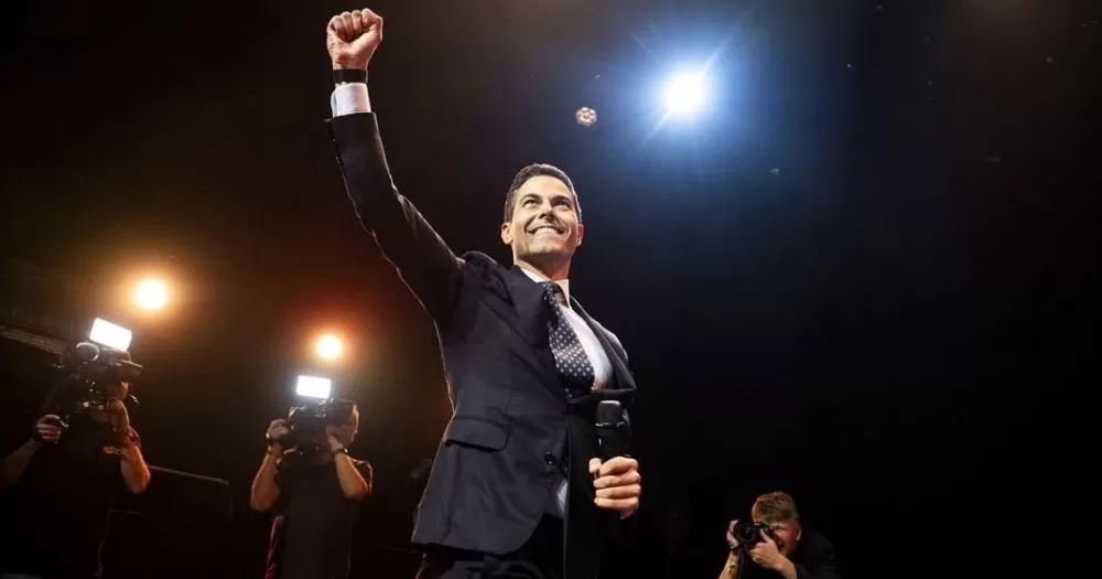 Rob Jetten, who became The Netherlands' first gay Prime Minister, holding a microphone on a stage with his fist raised in victory.
