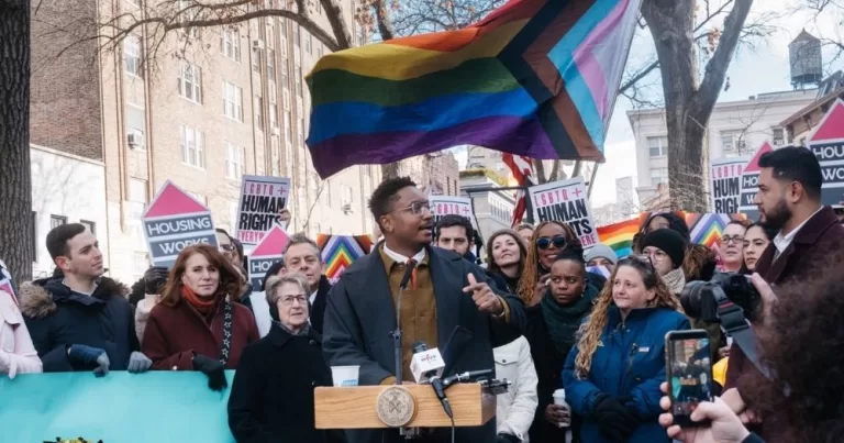 The image shows Council Member Chi Ossé standing at a platform at the Stonewall Monument and speaking, with a large pride flag in the background and a crowd gathered holding flags and placards.