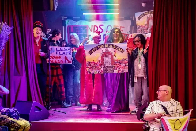 Revellers stand on stage in pink light at the Black Cap. 