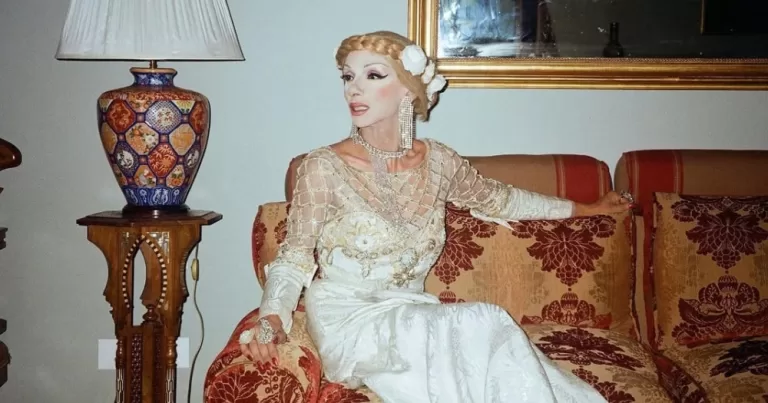 Artist Bassem Feghali dressed in a gown sitting on a couch.