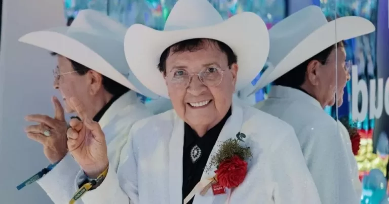 El Daña, the world's oldest drag king, smiles and poses in front of a large mirror.