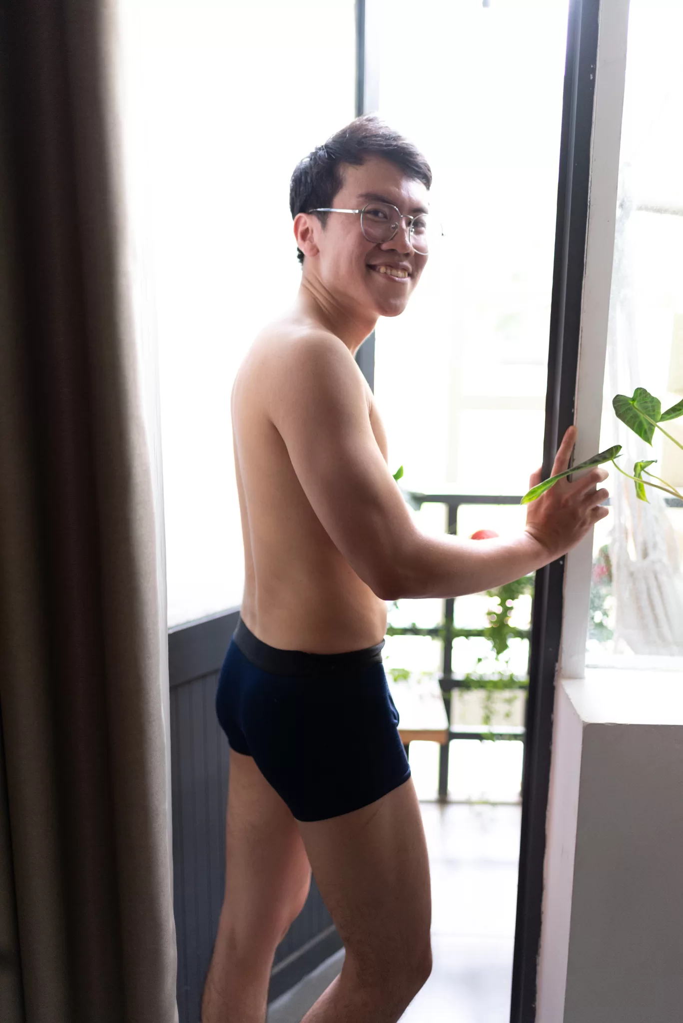 A young man poses in front of an open balcony door. He is in his underwear.