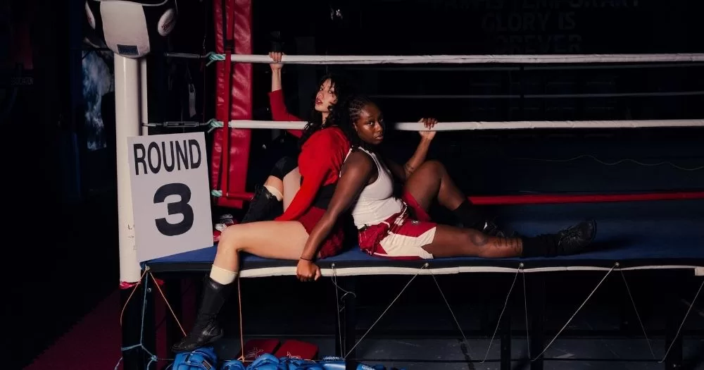 Two women, dressed as boxers sit back to back on the side of a boxing ring.