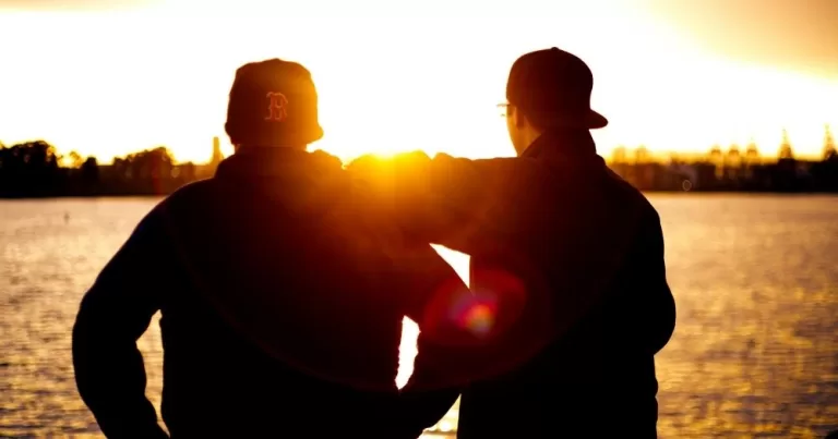 Two men stand together facing the sunset. This image represents the difficulty of navigating unrequited love and the idea of being 'out' in a friendship.