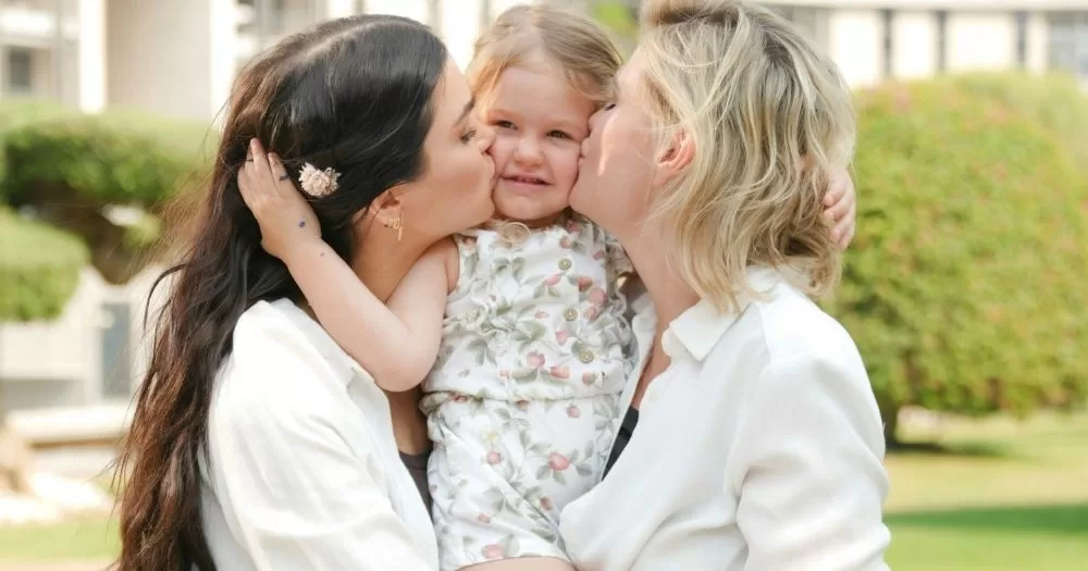 Two women hold a child. They are kissing her on the cheek. This image is being used to represent Irish legislation regarding LGBTQ+ families.