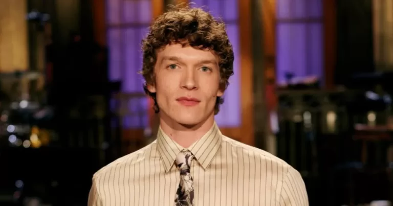 Connor Storrie posing on the studio stage in a light striped shirt and tie on SNL