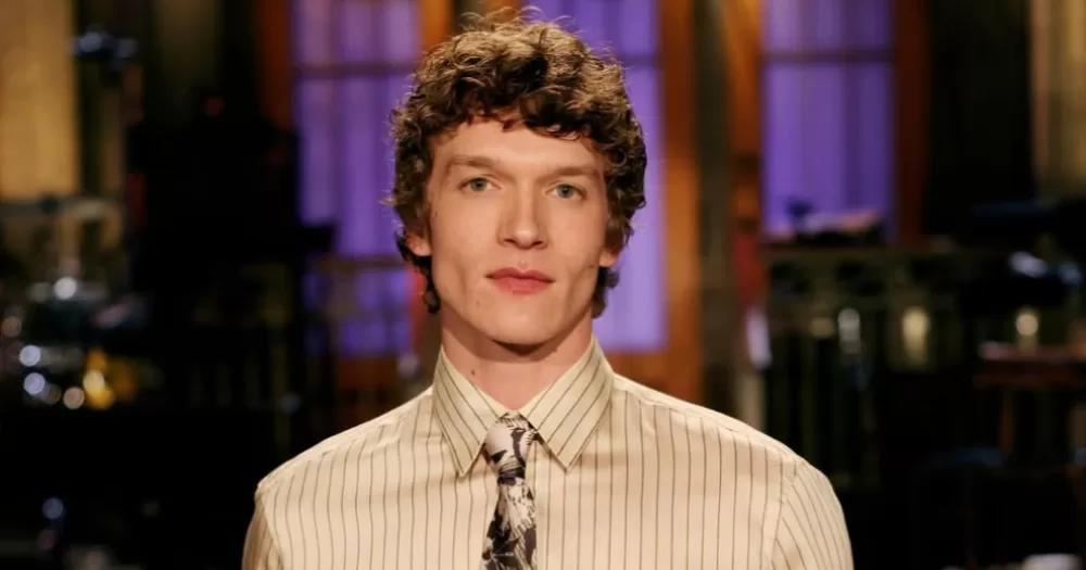 Connor Storrie posing on the studio stage in a light striped shirt and tie on SNL