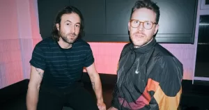 Music duo Phoeno pose in a kitchen, against a pink wall and a black cabinet.