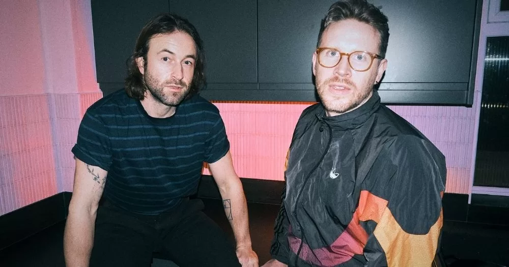 Music duo Phoeno pose in a kitchen, against a pink wall and a black cabinet.