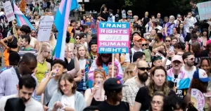 This article is about the NHS blocking new referrals for hormone treatment. In the photo, people marching carrying trans flags and banners that read 'trans rights are human rights'.
