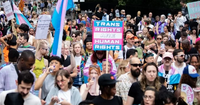 This article is about the NHS blocking new referrals for hormone treatment. In the photo, people marching carrying trans flags and banners that read 'trans rights are human rights'.