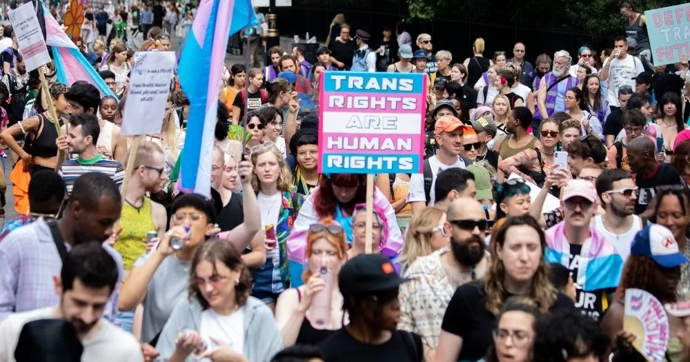 This article is about the NHS blocking new referrals for hormone treatment. In the photo, people marching carrying trans flags and banners that read 'trans rights are human rights'.