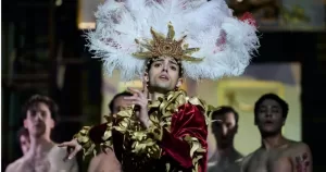 A dancer in the ballet Nureyev is mid-dance, wearing an elaborate costume and headpiece with several shirtless ballet dancers behind him.