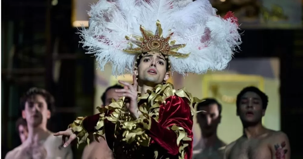A dancer in the ballet Nureyev is mid-dance, wearing an elaborate costume and headpiece with several shirtless ballet dancers behind him.