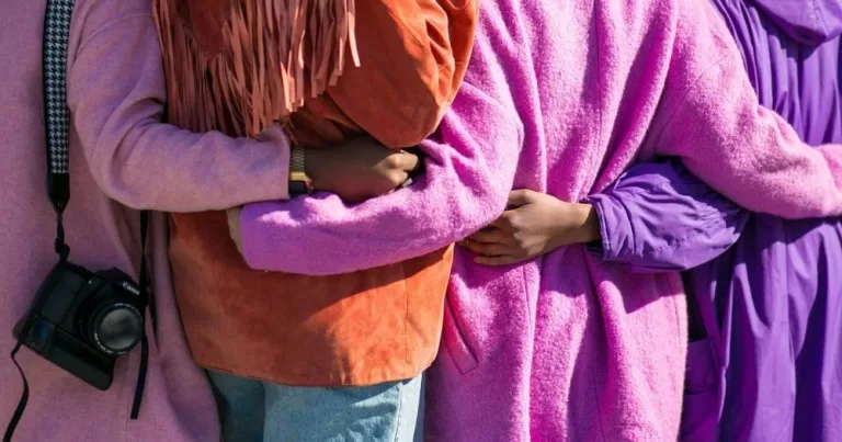 A group of people wearing colourful coats wrap their arms around one another. This image is being used to represent a queer PoC support group.