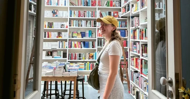 This article is about queer literature. In the photo, a girl smiling to the camera while standing in a library.