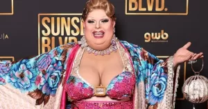 Maxi Shield, a drag queen, poses on a red carpet. She is wearing a colourful outfit.